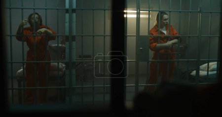 Photo for Two female prisoners in orange uniforms stand and walk behind bars in jail cell. Criminals serve imprisonment terms for crimes in prison. Women inmates in detention center or correctional facility. - Royalty Free Image