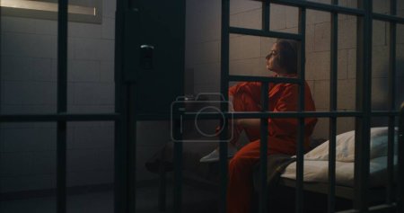 Téléchargez les photos : Prisonnière en uniforme orange donne le dîner du chariot de service de nourriture au criminel féminin dans la cellule de la prison. Femme criminelle assise sur le lit, purge une peine d'emprisonnement pour crime en prison ou dans un centre de détention. - en image libre de droit