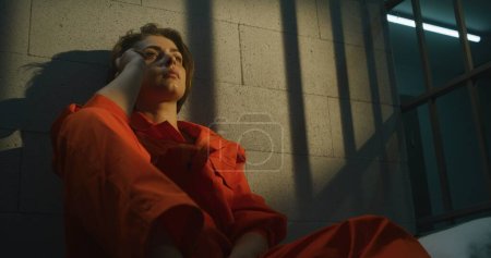 Photo for Woman criminal in orange uniform makes call, sits on bed in prison cell. Woman serves imprisonment term in jail. Detention center or correctional facility. - Royalty Free Image