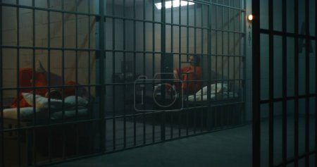 Foto de Una prisionera con uniforme naranja está detrás de las rejas de metal, otra se sienta en la cama en la celda de la prisión. Las mujeres cumplen penas de prisión por crímenes en la cárcel. Reclusos deprimidos en centro de detención. - Imagen libre de derechos
