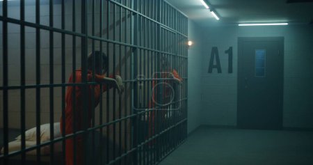 Eine Gefangene in orangefarbener Uniform steht hinter Metallgittern, eine andere sitzt auf dem Bett in der Gefängniszelle. Frauen verbüßen Haftstrafen für Verbrechen im Gefängnis. Depressive Häftlinge in Justizvollzugsanstalt.