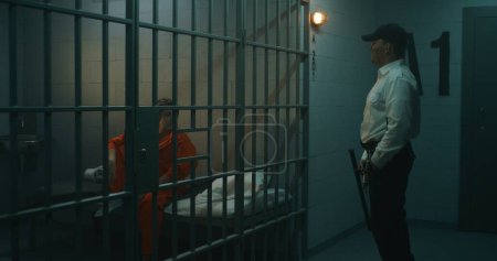 Foto de El oficial de la prisión camina por el pasillo, le da teléfono a una prisionera con uniforme naranja a través de barras de metal en la celda de la prisión. Las mujeres cumplen condena de prisión en prisión. Centro de detención o correccional. - Imagen libre de derechos