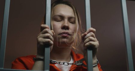 Photo for Female prisoner in orange uniform holds metal bars, stands in jail cell in handcuffs. Woman criminal serves imprisonment term for crime in prison. Depressed inmate in detention center. Close up. - Royalty Free Image