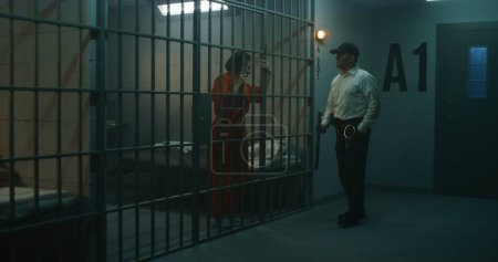 Jailer with police baton stands near prison cell, talks to female prisoner in orange uniform through metal bars. Women serve imprisonment terms in jail. Detention center or correctional facility.