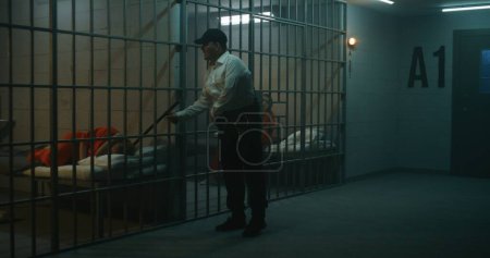 Foto de Carcelero con bastón de policía se para cerca de la celda de la prisión, habla con una prisionera con uniforme naranja a través de barras de metal. Las mujeres cumplen penas de prisión en prisión. Centro de detención o correccional. - Imagen libre de derechos