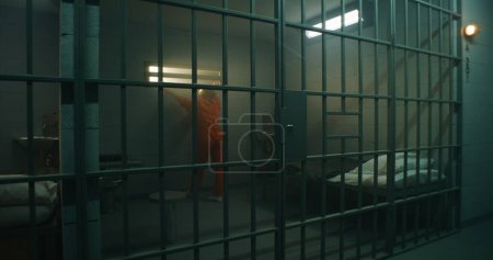 Foto de La prisionera de uniforme naranja camina y se para detrás de las rejas de metal, mira a la ventana cerrada en la celda de la cárcel. La mujer cumple condena de prisión por crimen en prisión. Deprimido criminal en centro de detención. - Imagen libre de derechos