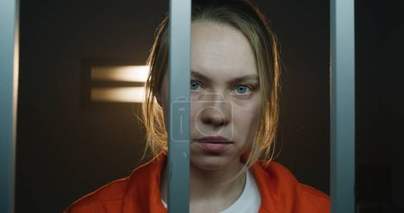 Photo for Scared female prisoner in orange uniform holds metal bars, stands in prison cell in handcuffs, looks at camera. Woman criminal serves imprisonment term for crime in jail or detention center. Portrait. - Royalty Free Image