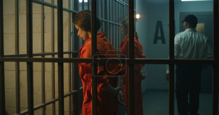 Téléchargez les photos : Deux prisonnières, détenues en uniforme orange, font face aux barreaux métalliques devant les cellules de la prison. Un officier de prison marche, regarde des femmes criminelles en prison. Centre de détention ou établissement correctionnel. - en image libre de droit