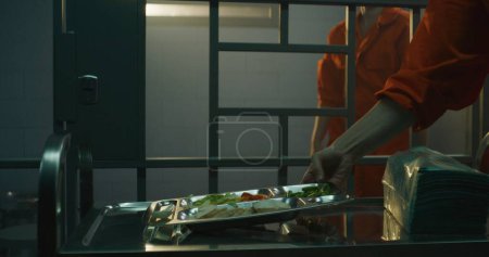 Foto de Trabajadora de la prisión da comida desde servir carro a prisionera con uniforme naranja en la celda de la cárcel. Una criminal se sienta en la cama, come la cena. Pena de prisión en prisión o centro de detención. Dolly disparó. - Imagen libre de derechos