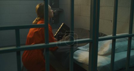 Foto de La prisionera de uniforme naranja se arrodilla cerca de la cama, ora a Dios en la celda de la prisión sosteniendo la Biblia. Mujer criminal cumple pena de prisión por delito en prisión, centro de detención o correccional. - Imagen libre de derechos