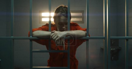 Photo for Female prisoner in orange uniform holds hands on metal bars, walks, looks at barred window in jail cell. Woman serves imprisonment term for crime in prison. Depressed inmate in correctional facility. - Royalty Free Image