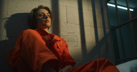 Téléchargez les photos : Une prisonnière en uniforme orange est assise sur son lit dans une cellule de prison, lit la Bible. Une femme criminelle purge une peine d'emprisonnement pour crime en prison ou dans un établissement correctionnel. - en image libre de droit