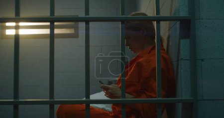 Téléchargez les photos : Prisonnière utilisant un smartphone en prison. Une femme en uniforme orange appelle, s'assoit sur le lit dans une cellule de prison. Il purge une peine d'emprisonnement. Centre de détention ou établissement correctionnel. - en image libre de droit