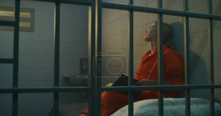 Téléchargez les photos : Une prisonnière en uniforme orange est assise sur un lit derrière des barreaux en métal, lit la Bible dans une cellule de prison, regarde une fenêtre barrée. Une femme criminelle purge une peine d'emprisonnement pour crime en prison ou dans un établissement correctionnel. - en image libre de droit