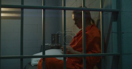 Foto de Prisionera usando smartphone en prisión. Mujer criminal con uniforme naranja hace una llamada, se sienta en la cama en la celda de la prisión. Cumplir condena a prisión en prisión. Centro de detención o correccional. - Imagen libre de derechos