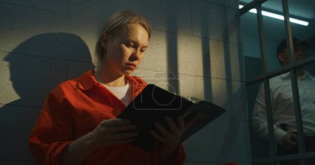 Foto de La prisionera de uniforme naranja se sienta en la cama en la celda de la prisión, lee la Biblia. El oficial de la prisión camina por el pasillo. Mujer criminal cumple pena de prisión por delito en prisión o correccional. - Imagen libre de derechos