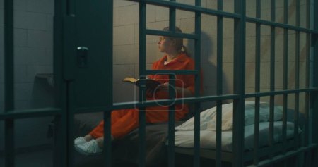 Foto de La prisionera de uniforme naranja se sienta en la cama detrás de las rejas de metal, lee la Biblia en la celda de la prisión, mira a la ventana con rejas. Mujer criminal cumple pena de prisión por delito en prisión o correccional. - Imagen libre de derechos