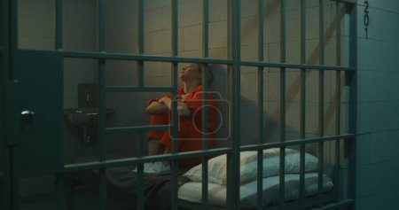 Foto de Prisionera de uniforme naranja sentada en la cama en la celda, mirando a la ventana cerrada. La mujer cumple condena de prisión por crimen en prisión. Deprimido criminal en centro de detención. - Imagen libre de derechos