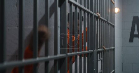 Foto de Un prisionero afroamericano con uniforme naranja se apoya en las rejas de las celdas. Vecino criminal sale de la mano de la puerta cerrada. Asesinos cumplen pena de prisión en correccional o centro de detención. - Imagen libre de derechos