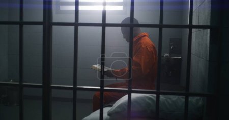 Photo for African American prisoner in orange uniform sits on the bed, reads Bible in prison cell. Male criminal serves imprisonment term for crime in jail or detention center. Faith in God. View through bars. - Royalty Free Image