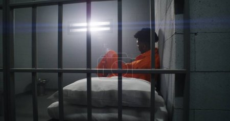 Foto de Dos presos vecinos cumplen condena de prisión en celdas de prisión. Un criminal afroamericano con uniforme naranja se sienta en la cama de la cárcel y mira a la ventana cerrada. Reclusos sombríos en el centro de detención. - Imagen libre de derechos
