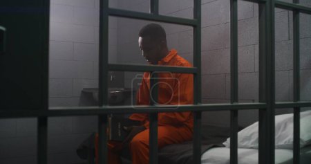 Photo for African American prisoner in orange uniform sits on the bed behind bars, reads Bible in prison cell. Criminal serves imprisonment term for crime in jail. Detention center or correctional facility. - Royalty Free Image