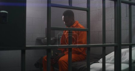 Un prisionero afroamericano con uniforme naranja se sienta en la cama tras las rejas, lee la Biblia en la celda de la prisión. Criminal cumple pena de prisión por crimen en la cárcel. Centro de detención o correccional.