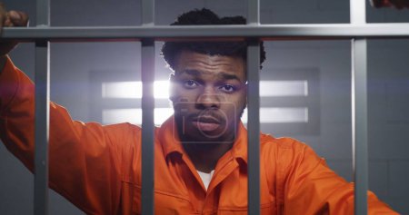 Foto de Hombre afroamericano en uniforme naranja mira a la cámara y amasa el cuello en la celda de la prisión. El prisionero cumple condena en prisión. Criminal en correccional o centro de detención. Vista de retrato. - Imagen libre de derechos