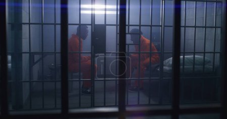Foto de Los prisioneros afroamericanos con uniformes naranjas se sientan en las camas de la cárcel y hablan emocionalmente. Un hombre toca a otro y empiezan a empujar. Los reclusos cumplen penas de prisión por delitos en el centro de detención. - Imagen libre de derechos