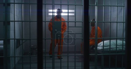 Foto de Dos prisioneros afroamericanos con uniformes naranjas hablan. Un hombre se sienta en la cama, otro entra en la celda de la cárcel. Hombres cumplen penas de prisión por delitos en la cárcel, centro de detención o correccional. - Imagen libre de derechos