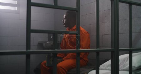 Téléchargez les photos : Prisonnier afro-américain en uniforme orange assis sur le lit derrière les barreaux, lit Bible en cellule de prison. Le criminel purge une peine d'emprisonnement pour crime en prison. Centre de détention ou établissement correctionnel. - en image libre de droit