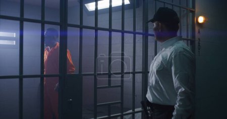 Prison employee puts on handcuffs on guilty inmate in orange uniform and leads him out of jail cell. Inmates serve imprisonment terms for crimes in prison, detention center or correctional facility.