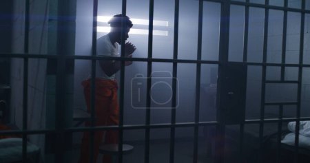 Foto de Un prisionero afroamericano con uniforme naranja hace boxeo en la celda. El carcelero camina por el pasillo de la prisión. Recluso cumple pena de prisión por delitos en centro de detención o correccional. - Imagen libre de derechos