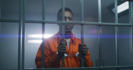 Téléchargez les photos : Un prisonnier afro-américain en uniforme orange garde les mains menottées sur les barreaux de prison et regarde la caméra. Un criminel coupable purge une peine d'emprisonnement. Triste meurtrier dans un établissement correctionnel. - en image libre de droit
