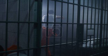 Male African American prisoner in orange uniform does shadow boxing in jail cell. Jailer walks prison corridor. Inmate serve imprisonment term for crimes in detention center or correctional facility.