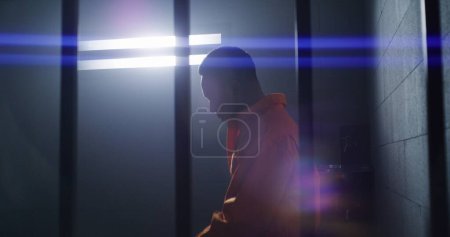 Téléchargez les photos : Un Afro-Américain déprimé en uniforme orange se tient debout du lit de prison, marche dans la cellule de prison et regarde la fenêtre barrée. Triste prisonnier purge une peine d'emprisonnement. Assassinat dans un établissement correctionnel sombre. - en image libre de droit
