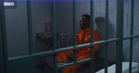 Foto de El hombre afroamericano se sienta en la cama y habla con el criminal en la celda de la prisión. Dos prisioneros con uniformes naranjas en correccional. Los reclusos cumplen penas de prisión por delitos en el centro de detención. - Imagen libre de derechos