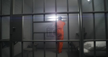 Foto de El prisionero afroamericano con uniforme naranja mira a la ventana cerrada y se apoya en las rejas de las celdas de la prisión. Reclusos deprimidos en el centro de detención. Asesinos cumplen pena de prisión en celda. - Imagen libre de derechos