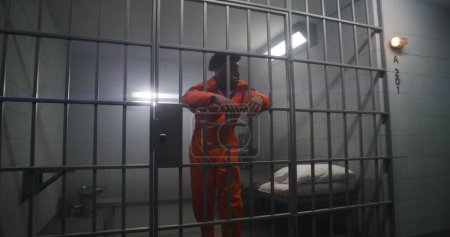 Téléchargez les photos : Un prisonnier afro-américain en uniforme orange se penche sur les barreaux de prison et regarde autour de lui. Un gangster coupable purge une peine d'emprisonnement dans un établissement correctionnel. Détenu sombre dans un centre de détention. - en image libre de droit