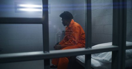 Foto de Reclusos afroamericanos en centros de detención o correccionales. Hombre deprimido en uniforme naranja se sienta en la cama de la prisión y mira a la ventana cerrada. Presos cumplen pena de prisión en celda. - Imagen libre de derechos