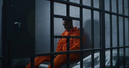 Foto de Un afroamericano culpable con uniforme naranja se sienta en la cama de la prisión y piensa en la libertad. Delincuente sombrío en correccional o centro de detención. Prisionero cumple pena de prisión en celda. - Imagen libre de derechos