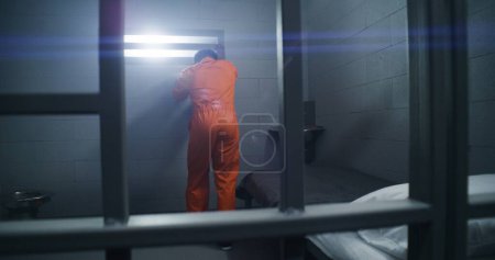 Téléchargez les photos : Un prisonnier afro-américain en uniforme orange se lève du lit et se penche sur les barreaux de la prison. Les criminels purgent une peine d'emprisonnement dans un établissement correctionnel. Des gangsters dans un centre de détention. - en image libre de droit