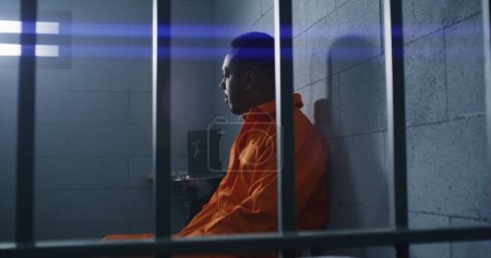 Foto de Un afroamericano culpable con uniforme naranja se sienta en la cama de la prisión y piensa en la libertad. Delincuente sombrío en correccional o centro de detención. Prisionero cumple pena de prisión en celda. - Imagen libre de derechos