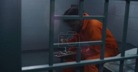 Foto de El hombre afroamericano se sienta en la cama, cena, cumple condena por crimen. El prisionero en uniforme naranja da comida de servir carro a criminal en la celda de la cárcel. Prisión o correccional. - Imagen libre de derechos
