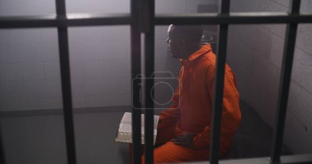 Téléchargez les photos : Un prisonnier afro-américain en uniforme orange est assis sur son lit derrière les barreaux, lit la Bible en cellule de prison. Un homme criminel purge une peine d'emprisonnement pour crime en prison. Centre de détention ou établissement correctionnel. - en image libre de droit