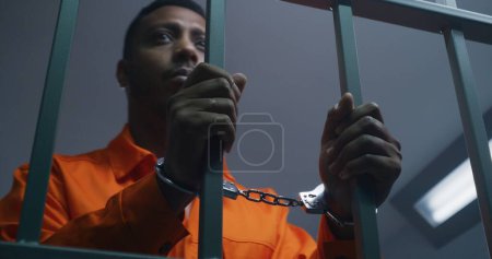 Foto de Hombre afroamericano con uniforme naranja mantiene las manos esposadas en las barras de celdas de la cárcel. Asesino deprimido cumple condena de prisión en prisión. Preso culpable en correccional o centro de detención. - Imagen libre de derechos