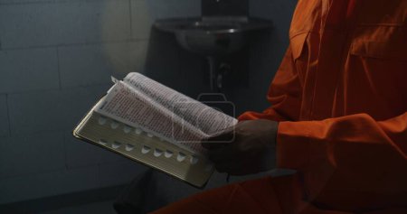 Photo for African American prisoner in orange uniform reads Bible in prison cell, turns pages. Male criminal, inmate serves imprisonment term for crime in jail or detention center. Concept of faith in God. - Royalty Free Image