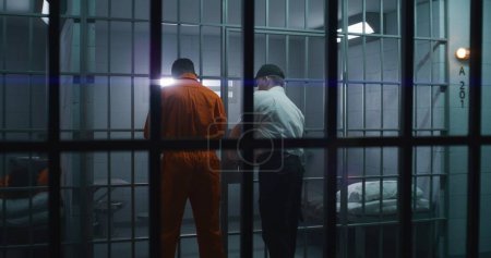 Photo for Warden brings guilty prisoner in jail cell and takes off handcuffs. African American man serves imprisonment term in correctional facility or detention center. Murderer in prison cell. - Royalty Free Image