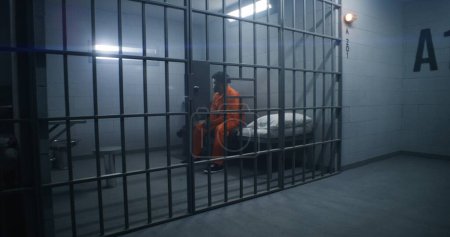 Téléchargez les photos : Détenus afro-américains dans un centre de détention ou un établissement correctionnel. Homme déprimé en uniforme orange assis sur le lit de la prison et regarde la fenêtre barrée. Les prisonniers purgent une peine d'emprisonnement en cellule. - en image libre de droit