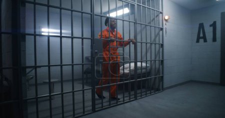 Photo for African American prisoner in orange uniform leans on jail cell bars and looks around. Guilty gangster serves imprisonment term in correctional facility. Gloomy inmate in detention center. - Royalty Free Image
